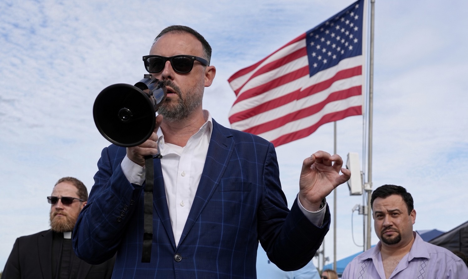 A man speaking into a megaphone beneath an American flag.